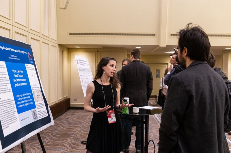 Sara, a white woman here in her 40s, is talking to people at a conference in front of a poster. She's wearing a black dress.