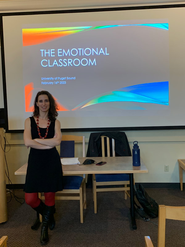 Sara (a fair-skinned brunette wearing a black dress and a bright red necklace and red tights) stands in front of a powerpoint slide that reads "The Emotional Classroom".